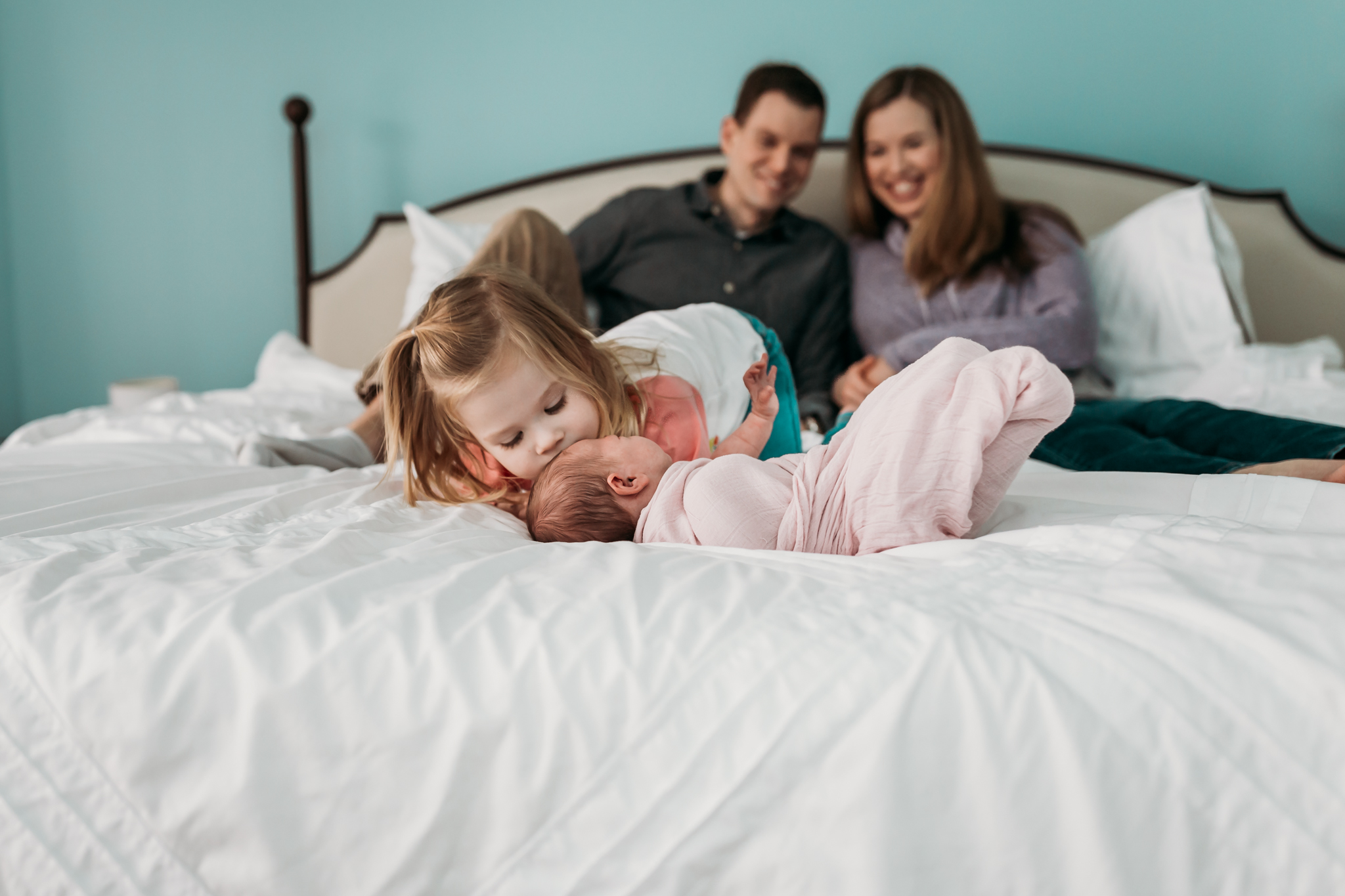 sister kisses newborn on bed while parents look on