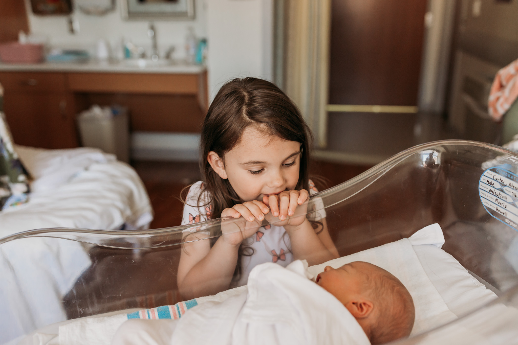 sister looks lovingly at newborn brother in bassinet
