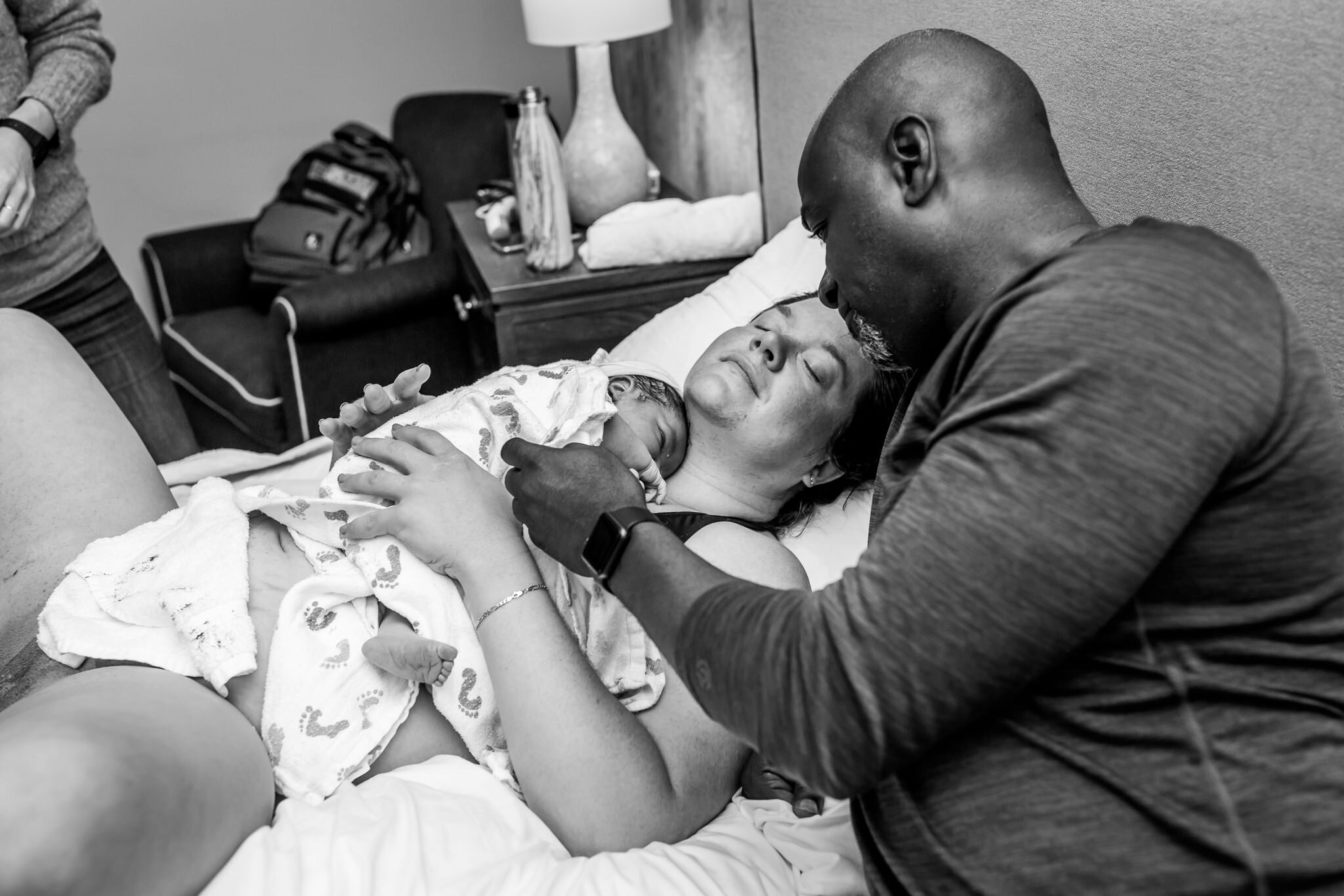 husband kisses wife as newborn lays on her chest postpartum