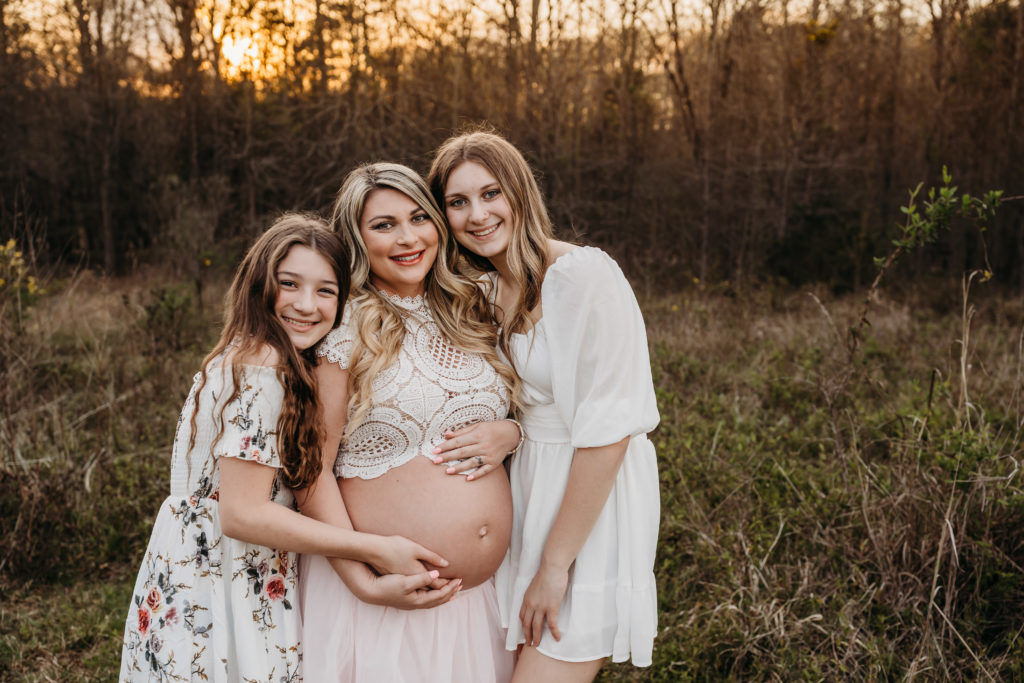 Pregnant mom in pink tulle skirt hugging her daughters in a field