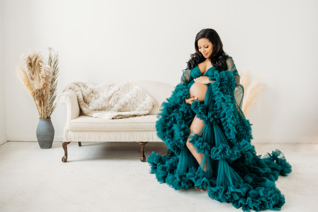 Woman looking down at exposed pregnant belly in teal tulle dress during studio maternity photoshoot