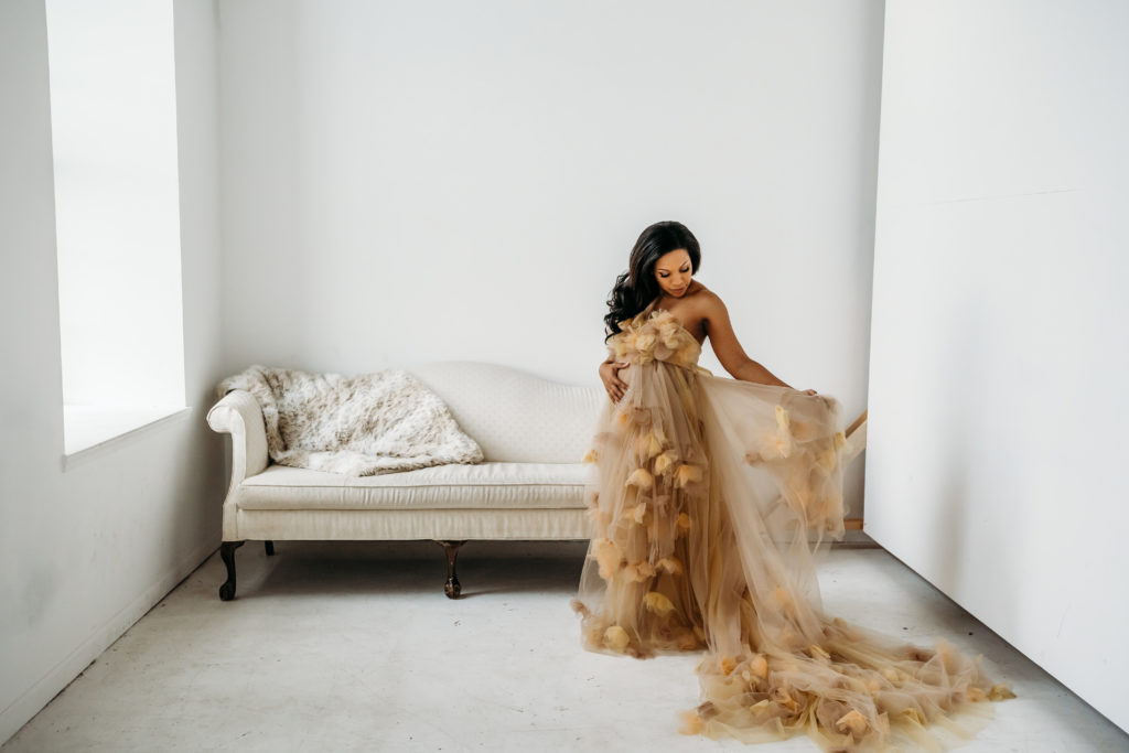 Pregnant woman standing in front of white couch in cream tulle dress looking at her dress during maternity studio session