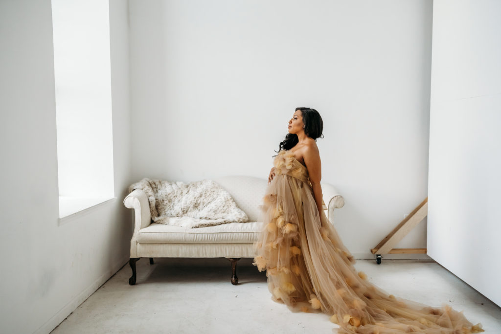 Pregnant woman standing in front of white couch in cream tulle dress looking out window during maternity studio session