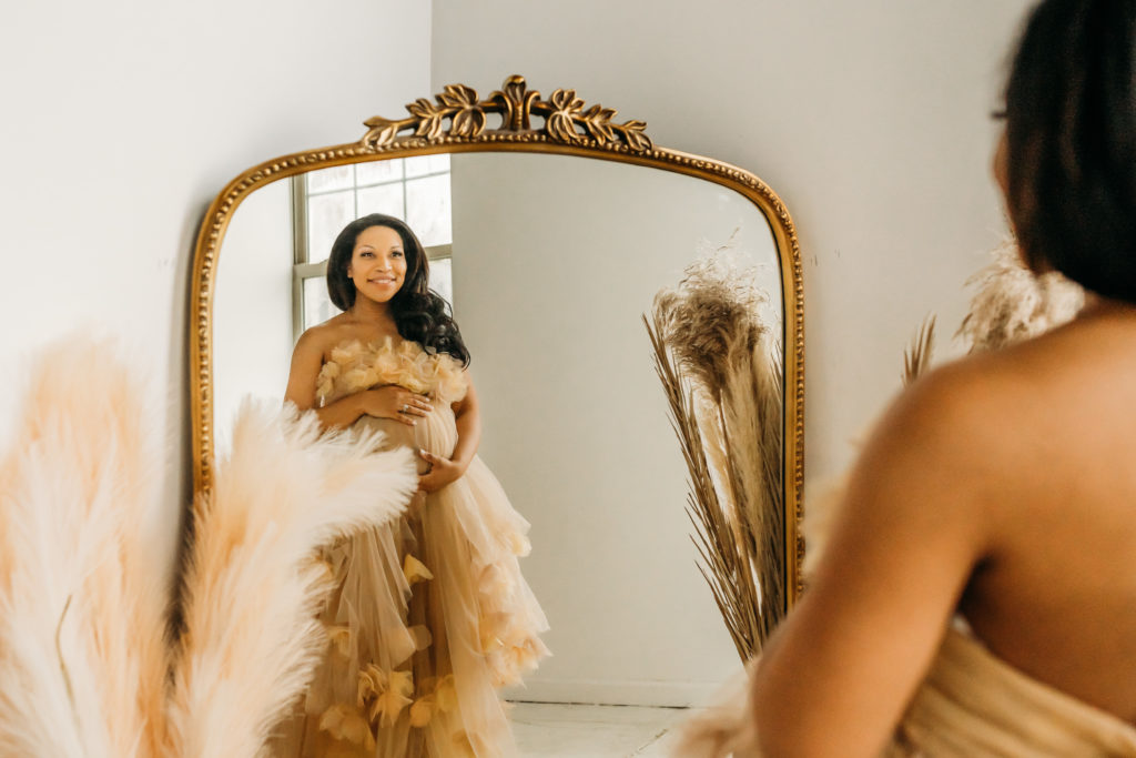 pregnant mom looking at herself in the mirror while wearing a cream tulle maternity dress