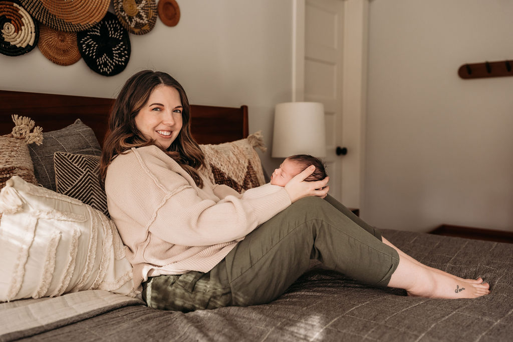 mom looking at camera while holding baby on bed during lifestyle newborn session
