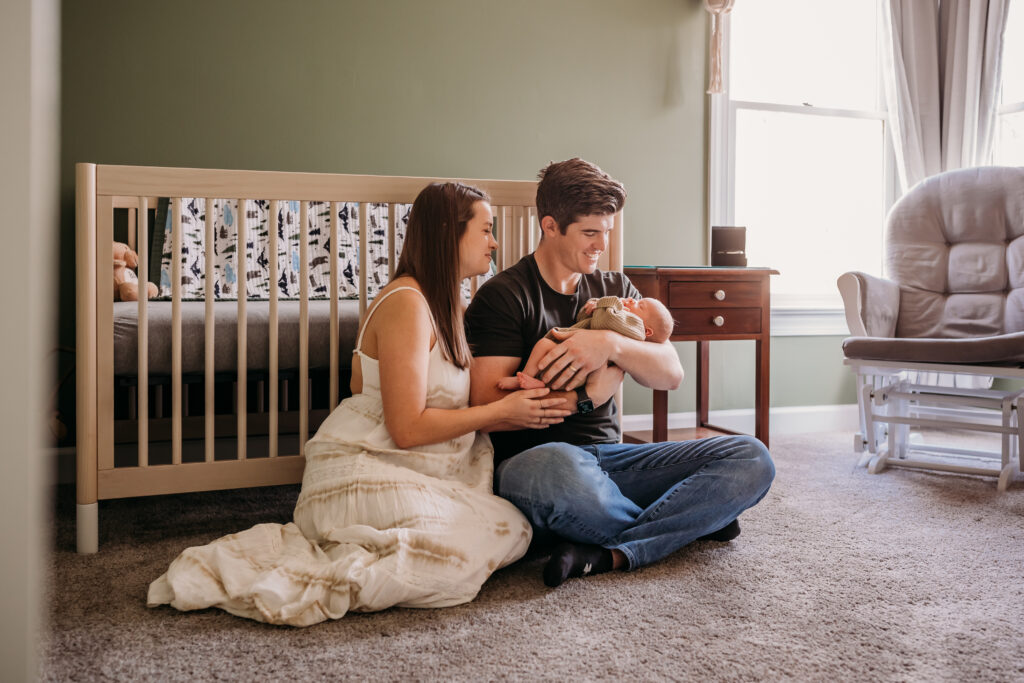 new parents hold baby while sitting next to crib