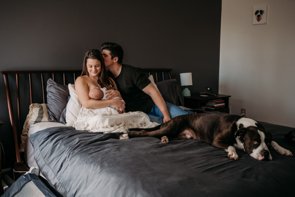 new mom breastfeeding newborn in bed while her husband kisses her and dog lays by her feet