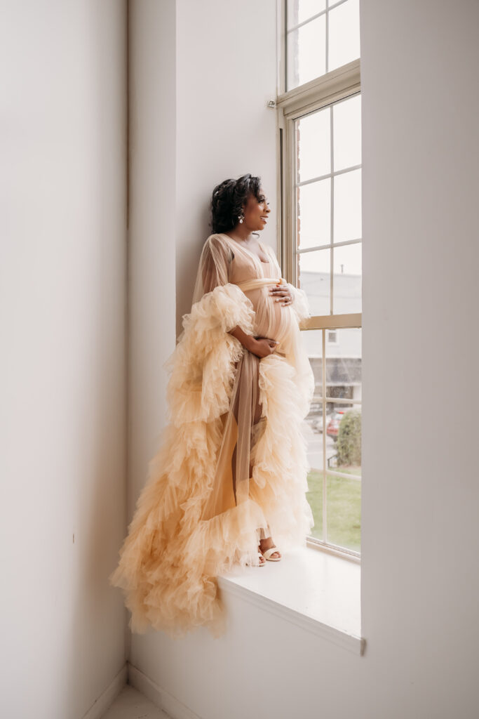 pregnant mother standing on window sill in cream tulle dress for maternity pregnancy cravings photoshoot