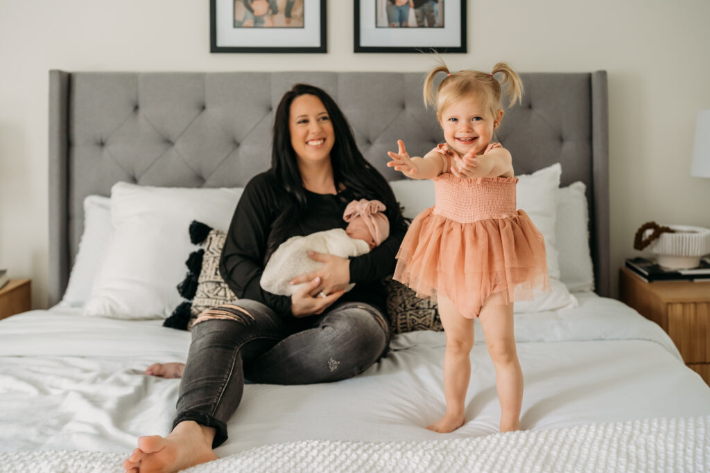 little girl stands with mom holding a baby on bed